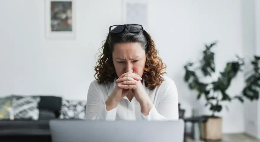 En kvinna med rufsigt hår och glasögon på huvudet sitter framför en laptop, djupt försjunken i tankar. Hennes ansiktsuttryck tyder på oro, kanske efter att ha utsatts för psykologisk misshandel.
