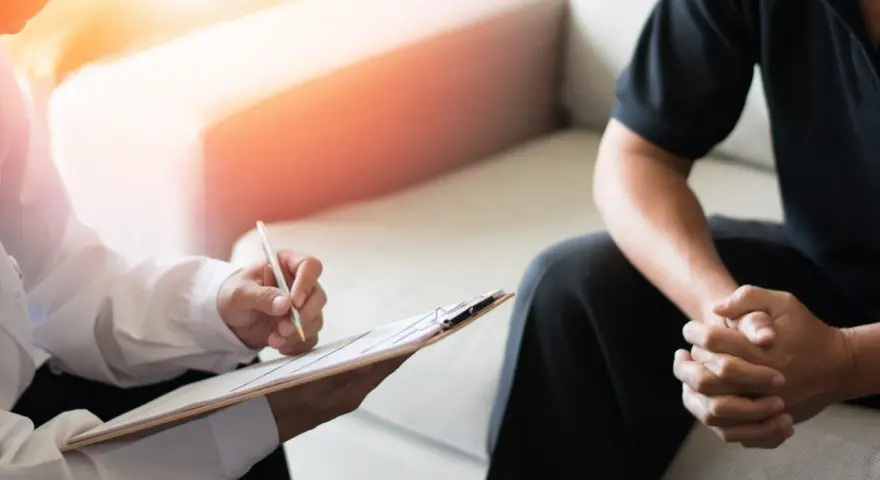 En terapeut skriver anteckningar medan en patient sitter med händerna knäppta. Bilden illustrerar vad betyder terapi – en plats för samtal och stöd.
