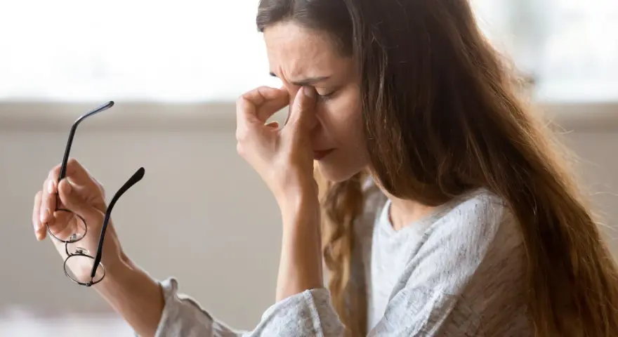 Kvinna med glasögon i handen, gnuggar sig i ögonen. Kanske tecken på muskelryckningar stress.