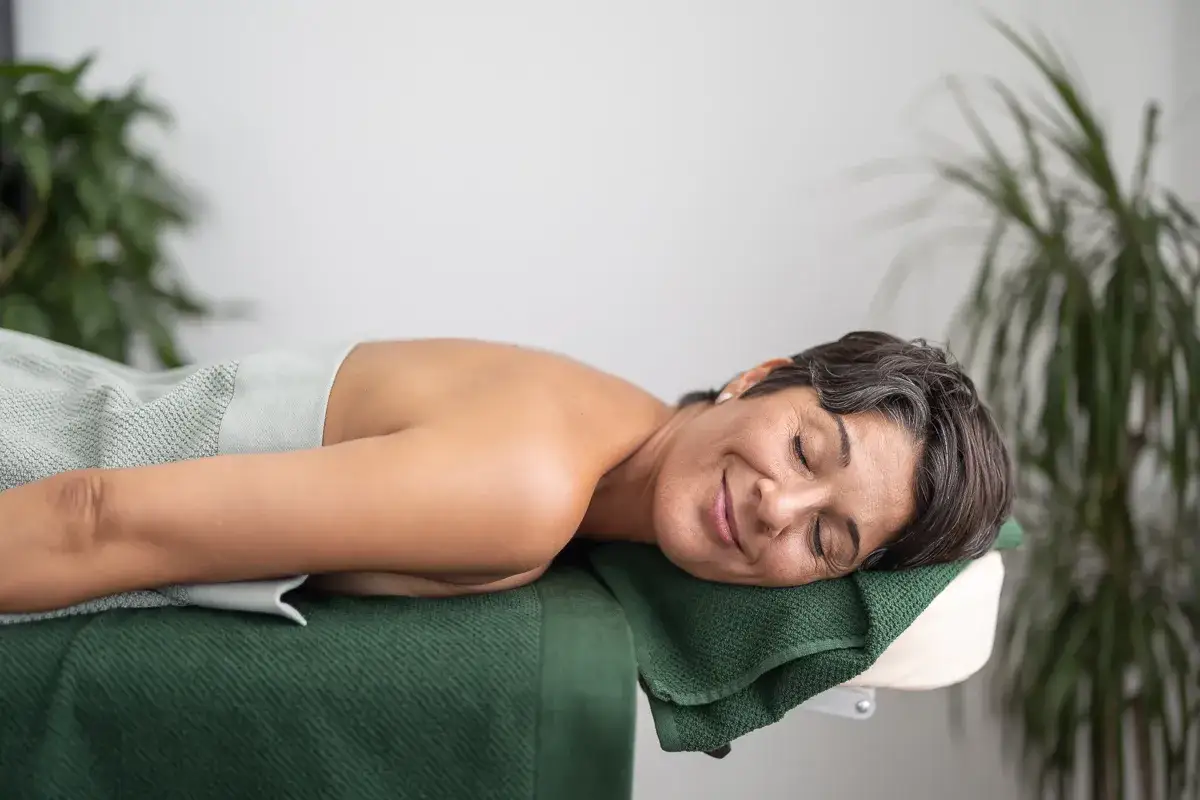 En kvinna vilar på en massagebänk, med slutna ögon och ett leende. Hon verkar finna ro och lindring från emotionell utmattning.