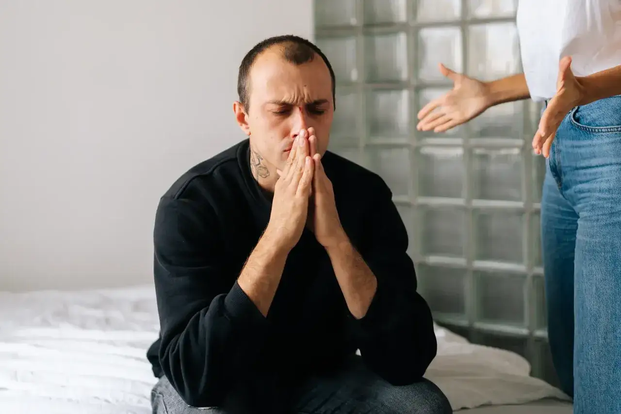 En man sitter med händerna knäppta och blicken nedåt, uppenbart ledsen. En kvinna står bakom honom och gestikulerar, vilket antyder en destruktiv relation.