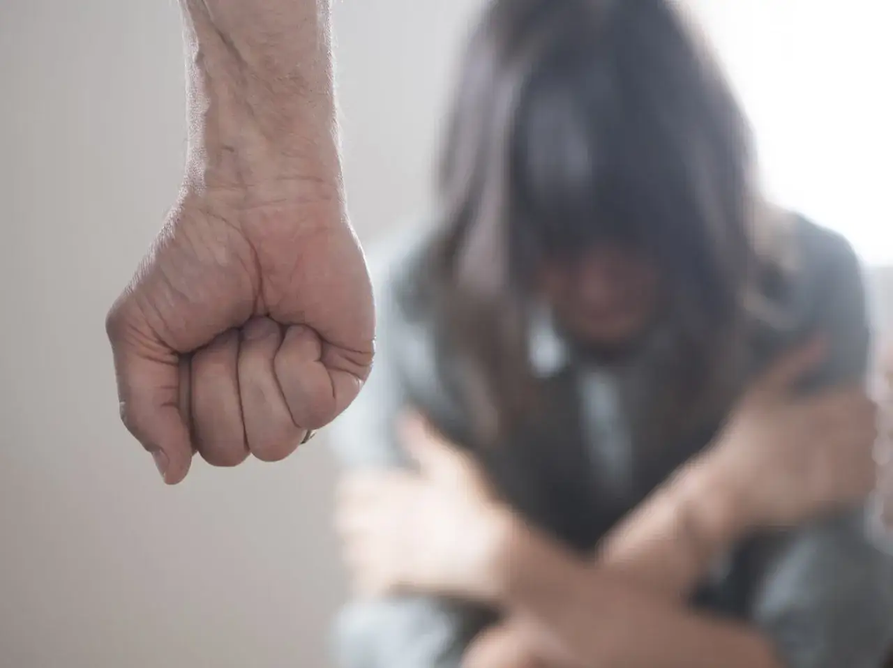 En knuten näve hotar en person som sitter hopkrupen. Bilden symboliserar fysisk misshandel och dess skrämmande konsekvenser.