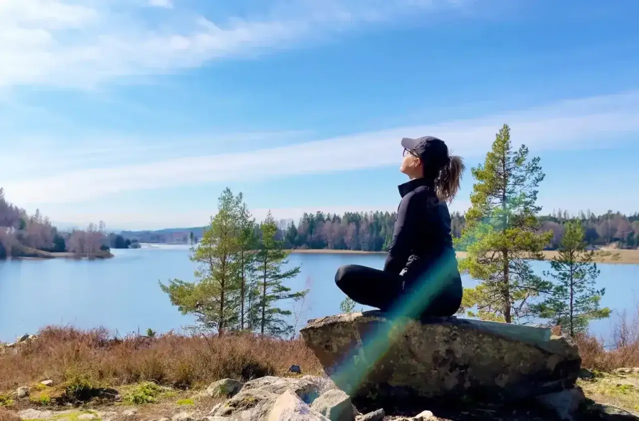 Kvinna sitter p&aring; en sten vid en sj&ouml; och samlar mental styrka. Solen lyser och himlen &auml;r bl&aring;.
