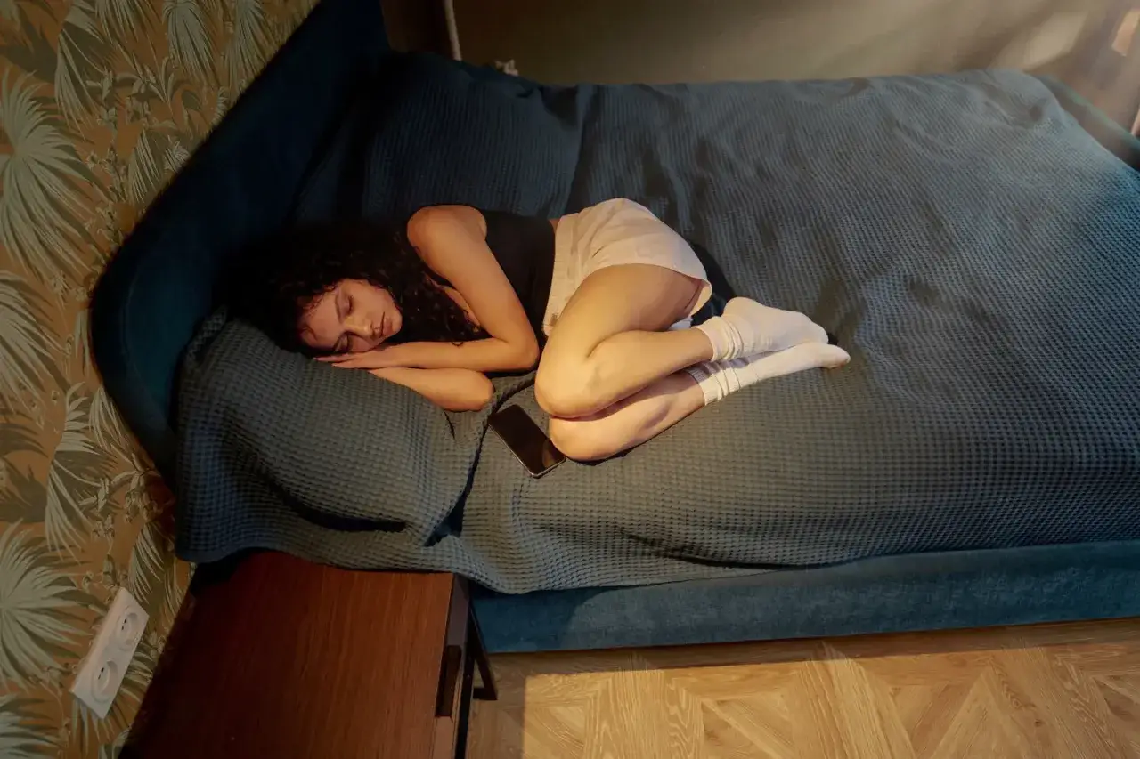 En kvinna sover på en säng, en mobiltelefon ligger bredvid henne. En bild av avslappning mot ångest.