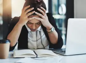 En kvinna med händerna i håret sitter vid ett skrivbord med en öppen anteckningsbok och en laptop. Hon ser stressad ut, vilket påverkar trivseln på jobbet.
