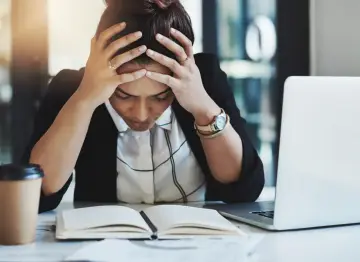 En kvinna med händerna i håret sitter vid ett skrivbord med en öppen anteckningsbok och en laptop. Hon ser stressad ut, vilket påverkar trivseln på jobbet.