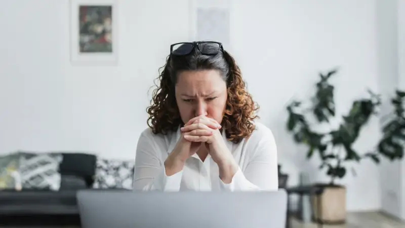 En kvinna med rufsigt hår och glasögon på huvudet sitter framför en laptop, djupt försjunken i tankar. Hennes ansiktsuttryck tyder på oro, kanske efter att ha utsatts för psykologisk misshandel.